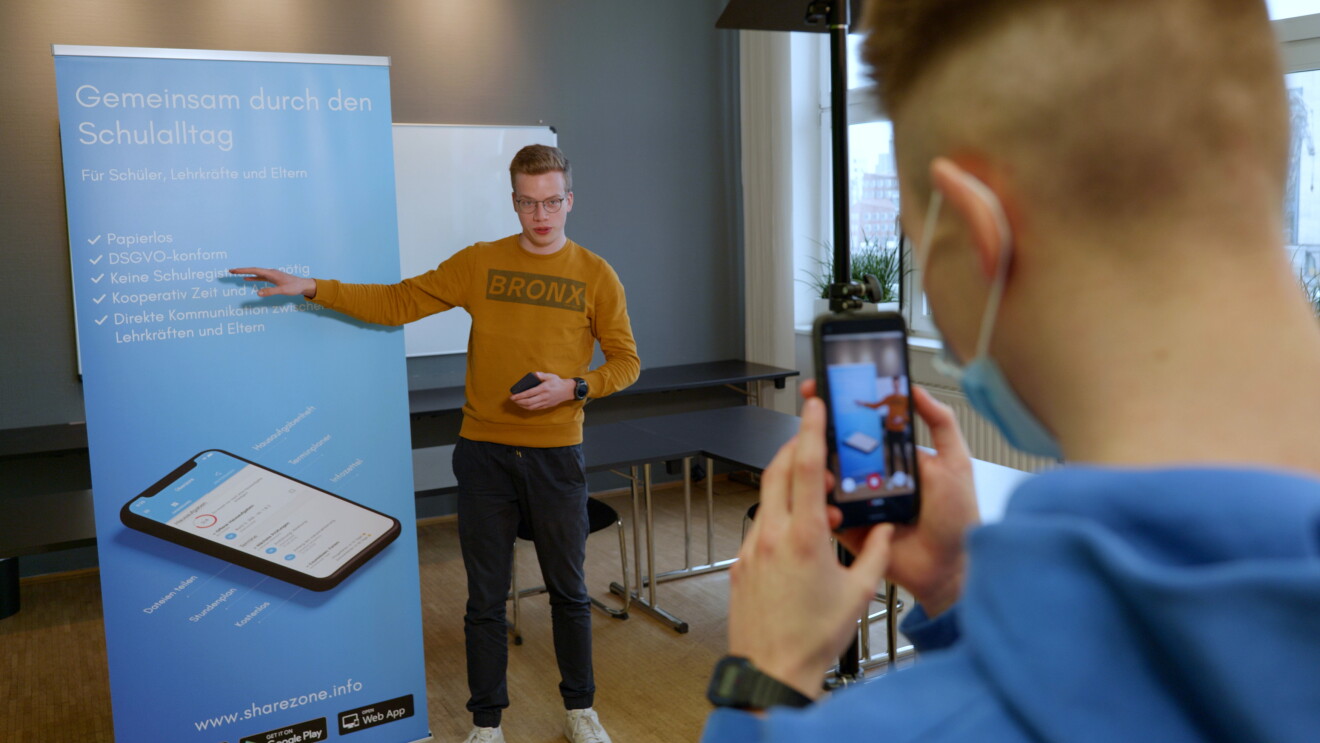Ein Mann mit orangenem Pulli steht vor einem Banner, der die App Sharezone präsentiert.