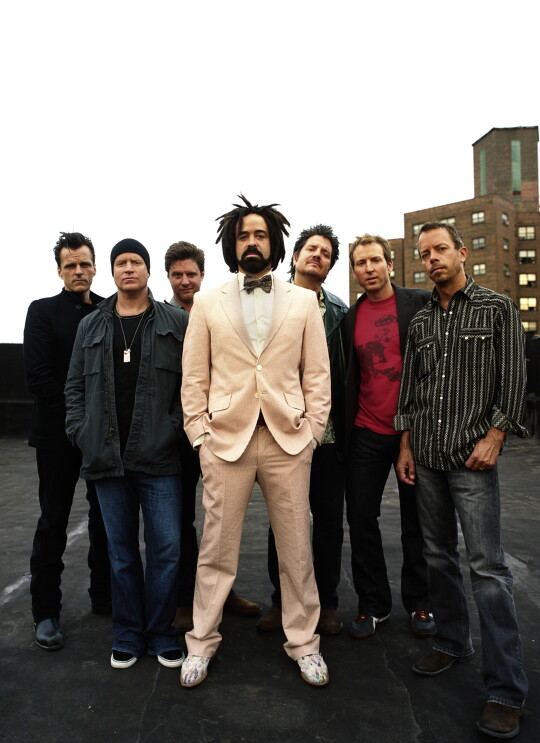 The Counting Crows band, six men stand together wearing casual clothing. In front of them, Adam Duritz stands in a cream suit with bow tie and tennis shoes.