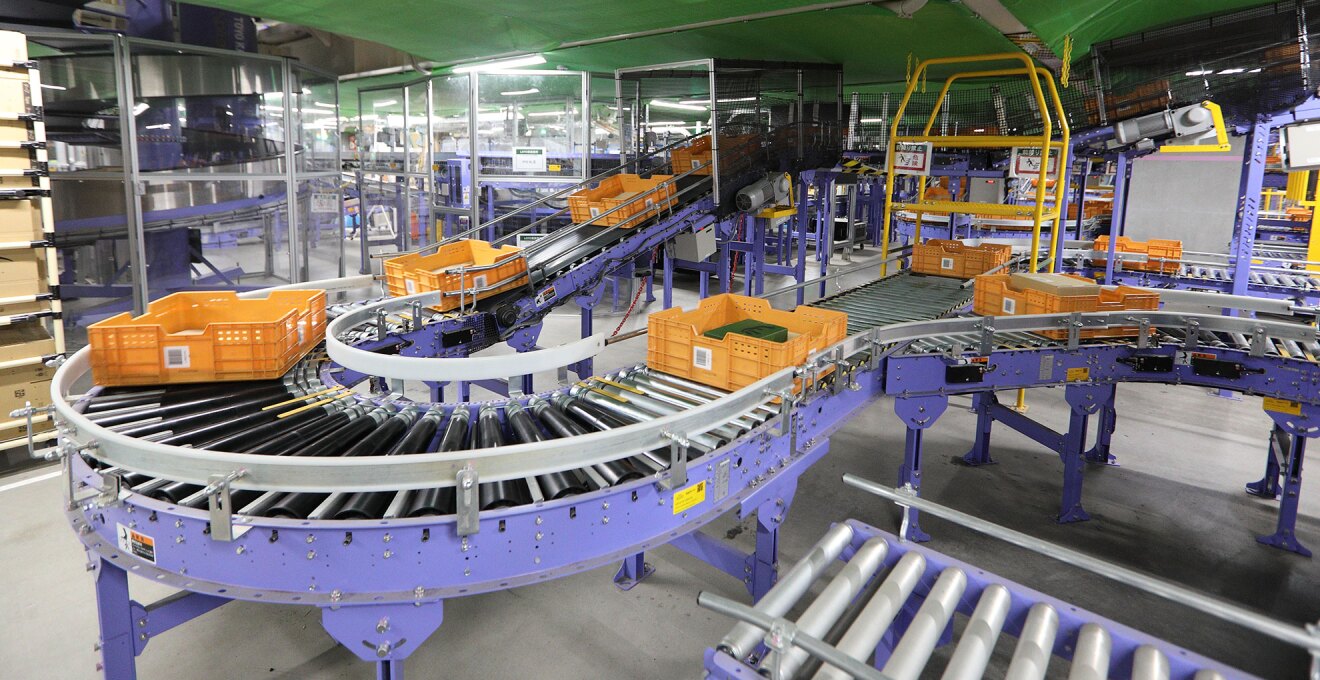 広大な物流センターの自動仕分けライン。側面と脚が紫色のコンベアベルトのレーン上を、オレンジ色のコンテナがいくつも流れていく。床はグレー、天井は緑色。
