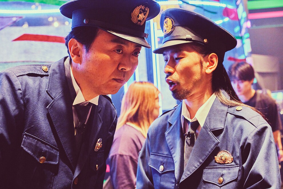 派手で明るいセットを背景に、警察官の制服衣装を着た2人の男性の上半身。右側の男性が話しかけ、左側の男性が耳を傾けている。2人の背景に複数のスタッフ。