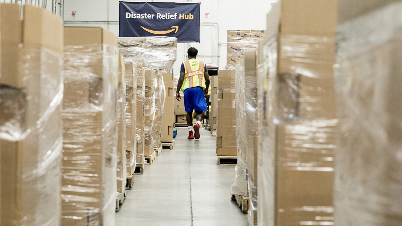 A man wearing shorts, tennis shoes, a t-shirt and safety vest walks between pallets of supplies in Amazon's Disaster Relief Hub