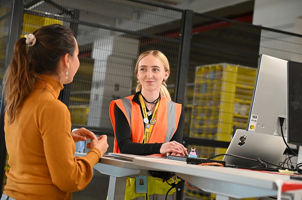 Logistikmitarbeiterin steht vor ihrem Schreibtisch und Laptop mit orangener Veste und redet mit einer Mitarbeiterin die zu Besuch ist.