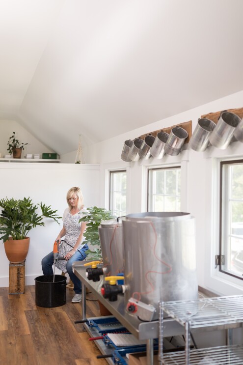 A woman mixes a small batch of soap with a power-mixer. In the foreground, are tanks used to heat and melt materials for the soaps and candles to be sold. Above her are several empty pots used for production.