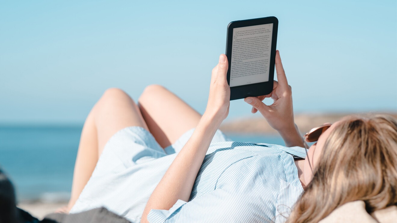 An image of a person laying down at the beach reading on their Amazon Kindle device and wearing a white and blue stripe dress. 