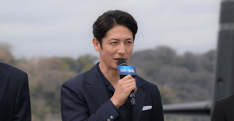 日中の屋外で、紺色のジャケットを着てマイクを持った男性。口にマイクを当て話している。背景に木々と黒い建造物のようなものが見える。