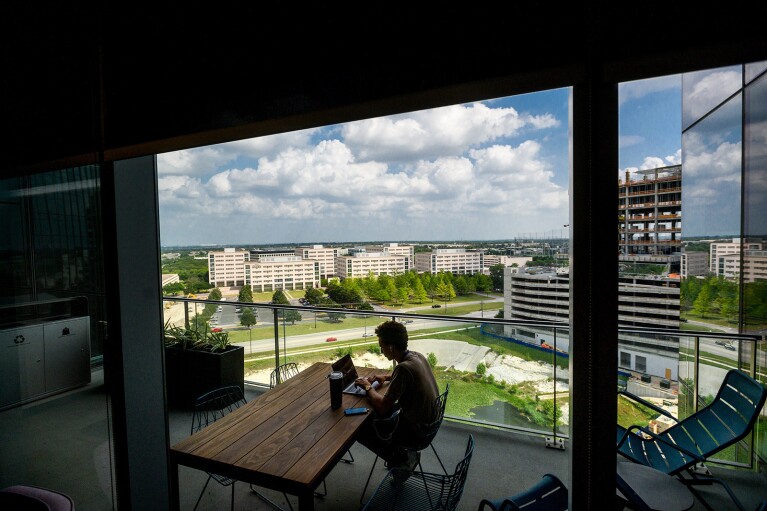 A man sits at a table near a window overlooking a green area and several other buildings.