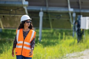 A woman wears an orange safey vest, a hard hat, and sungalsses as she walks outside by a solar farm.