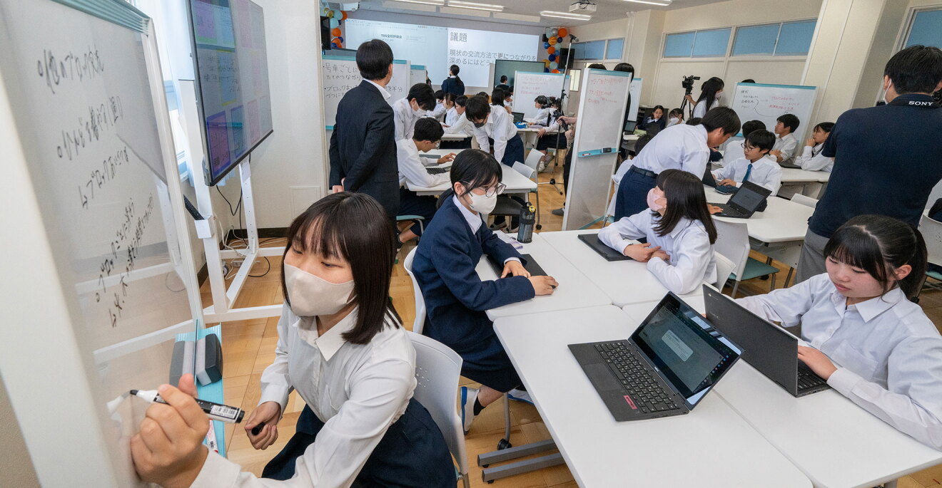 教室で制服の生徒たちが机をまとめた複数の班に分かれて話し合いをしている。各自机の上にノートパソコンがあり、一部の生徒は開いて使用している。班ごとに移動式ホワイトボードがあり、手前の生徒が黒いペンでボードに書き込んでいる。奥の壁に投影された画像には「議題」の字が読める。