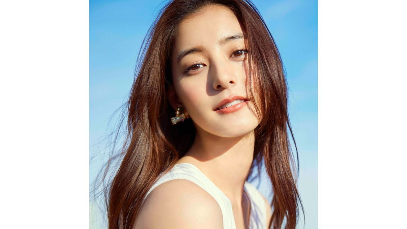 長い茶色の髪をしている女性(新木優子さん)が正面を向いて微笑んでいる。彼女は白いタンクトップを着て、体は横を向いている。