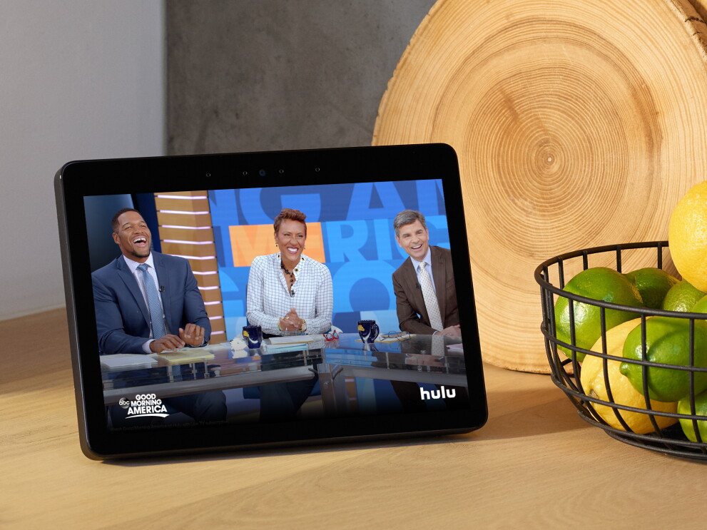 An Echo Show device on a kitchen counter, with Good Morning America displayed on the screen. To the right of the device is a cutting board made of a slice of wood, and a metal basket filled with lemons and limes. 