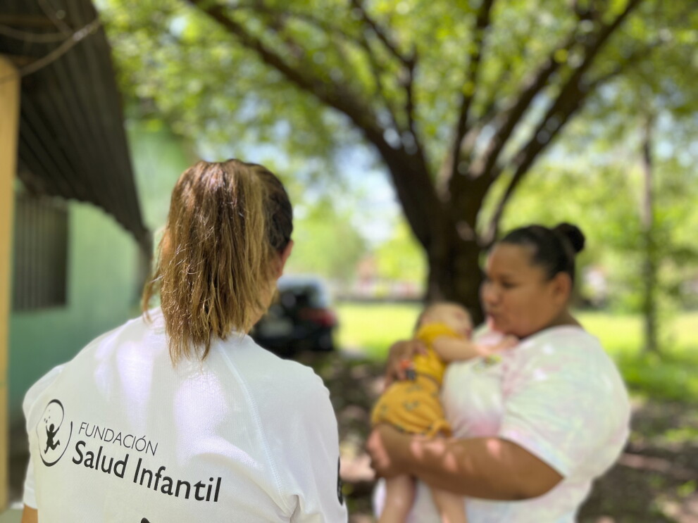 Cooperante de la Fundación Salud Infantil con una camiseta de la entidad en primer plano. De fondo una madre con su bebé en brazo. 