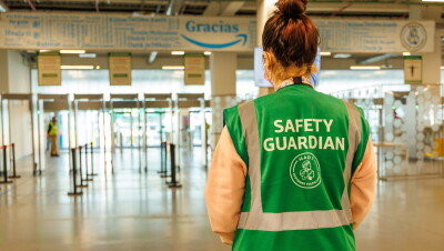 En la entrada de un centro logístico de Amazon aparece un Safety Guardian de espaldas. Es una mujer con una coleta recogida alta. La persona lleva mascarilla y un colar del que lleva el badge colgado. Tiene un chaleco de color de color verde con las letras blancas Safety Guardian MAD7.  