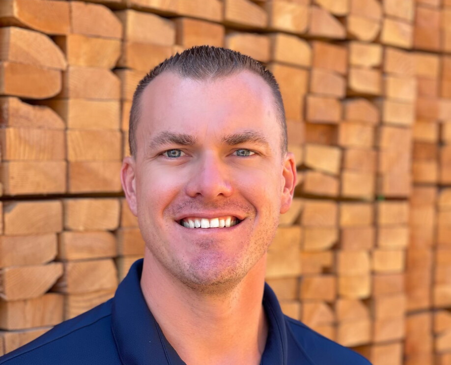 An image of a man smiling for a headshot photo. In the background there are stacks of wood pellets piled high.