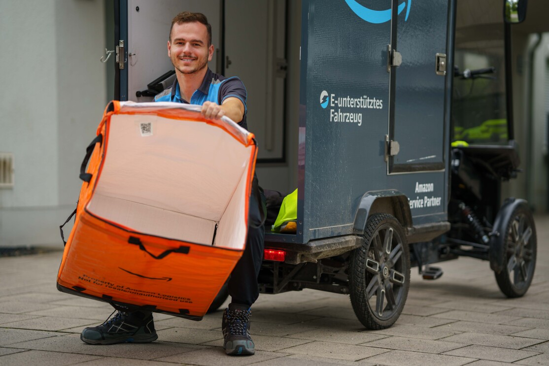 Enis sitzend auf der Transportfläche des E-Cargo-Bikes: Er hält eine große leere orangene Versandtasche vor sich, auf der ein großes Amazon-Logo zu erkennen ist.