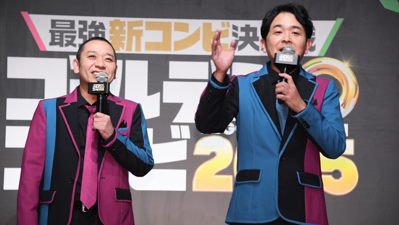 「最強新コンビ」などの字が書かれた黒壁を背に、黒・青・紅色の衣装を着た2人の男性がマイクを手に並んで立つ。