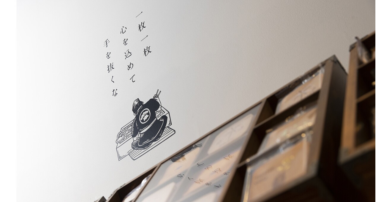 白い壁に「一枚一枚心を込めて手を抜くな」という言葉と、煎餅を焼く職人さんの背中が描かれている。