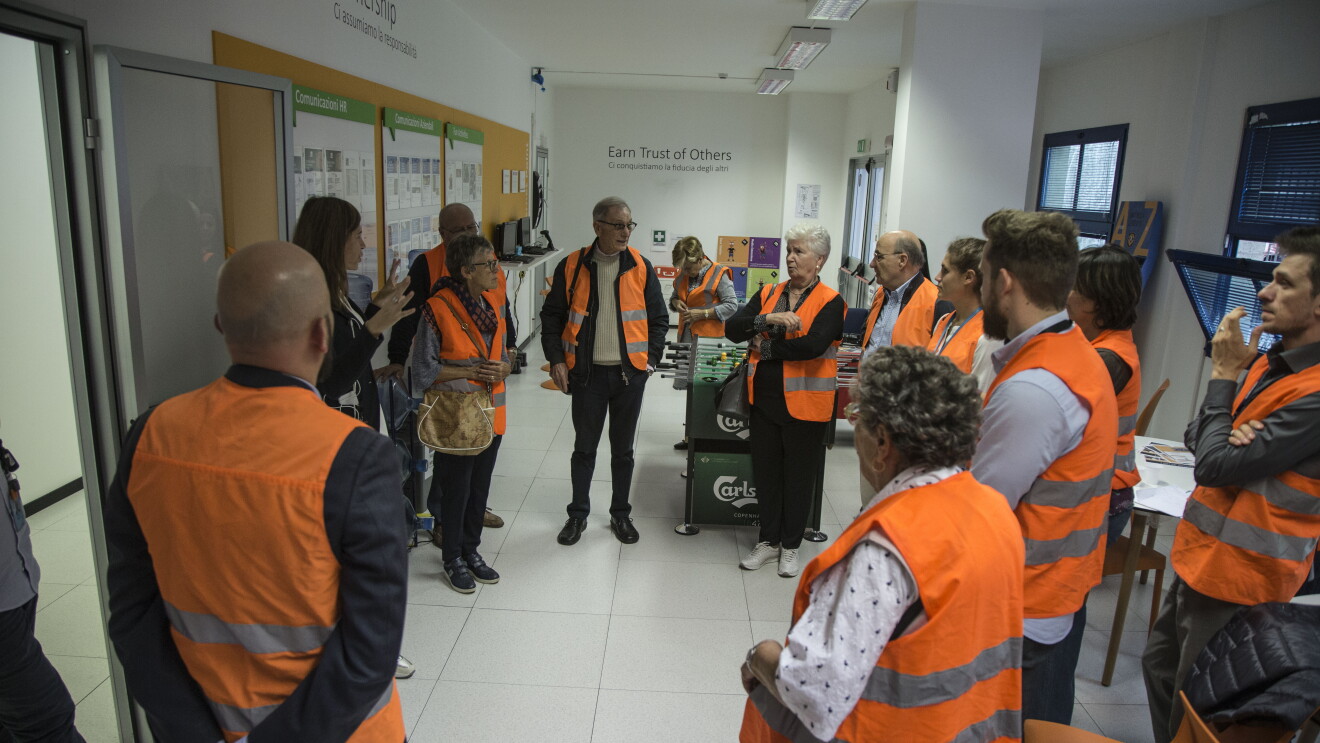Gruppo di nonni di dipendenti Amazon in visita al Centro di distribuzione urbana Prime Now di Milano in occasione della Festa dei nonni. Indossanno giubbini catarinfrangenti arancioni. Sui muri della sala in cui si trovano si leggono due principi di leadership di Amazon: 'Earn trust of others' e 'Ownership'