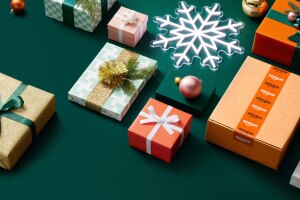 an image with a dark green background. There are various wrapped gifts in colorful packaging with ribbons, an Amazon box, and a light-up snowflake.