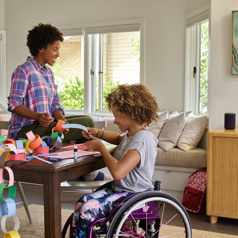 A mother sits on a dining room table as she and her daughter laugh while making a paper chain. Behind them, Amazon Echo is on a console table.