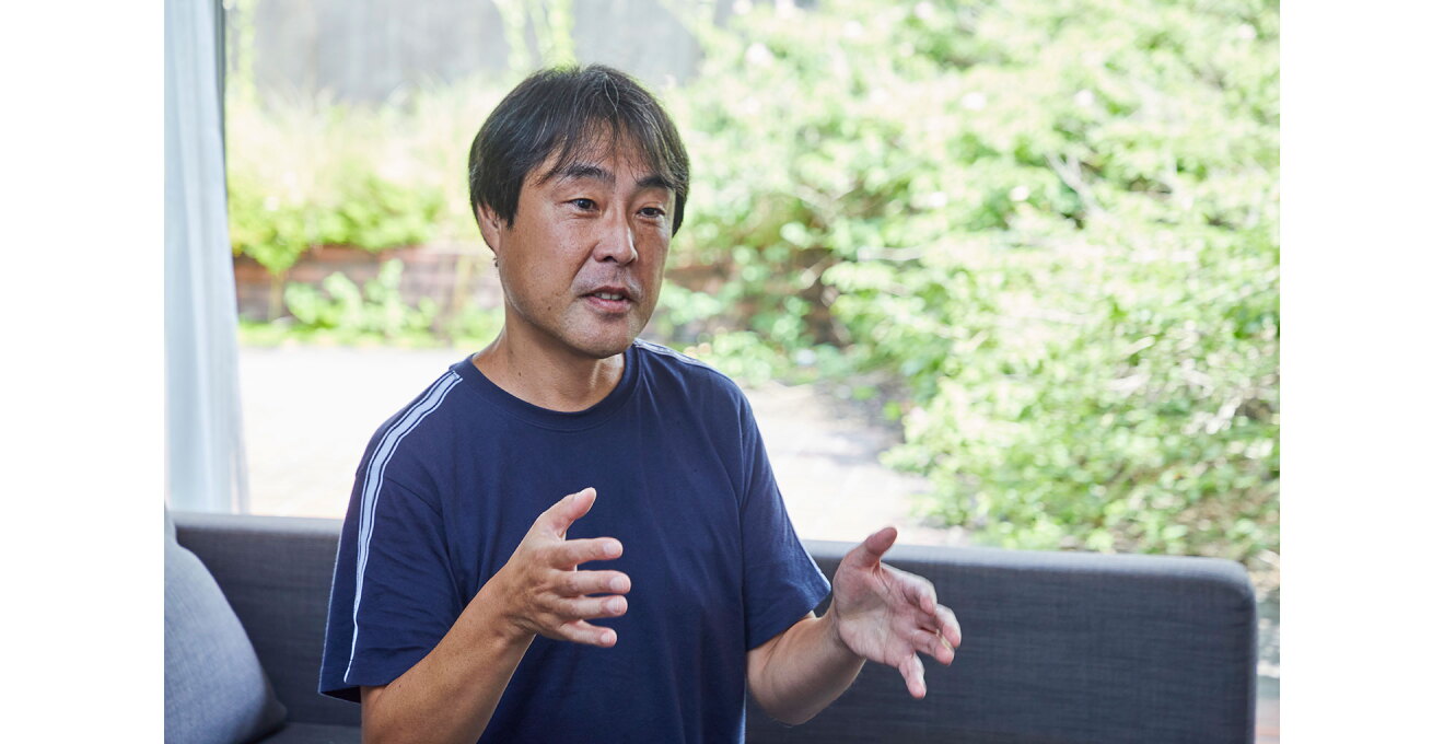 大きな窓を背に、ソファに座って身振り手振りをつけながら話をする人物。窓の向こうには緑が見える。