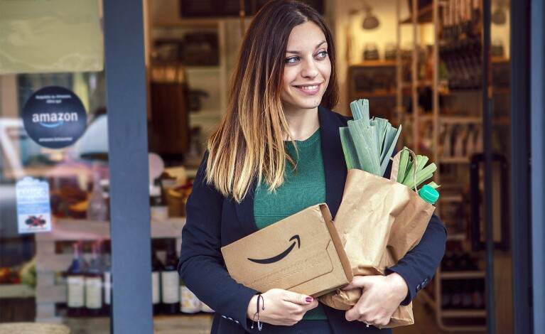 Eine junge Frau mit blonden langen Haaren beim Verlassen eines Geschäfts. Sie hält ein Amazon Päckchen und eine Papiereinkaufstüte mit Lebensmitteleinkäufen in der Hand.