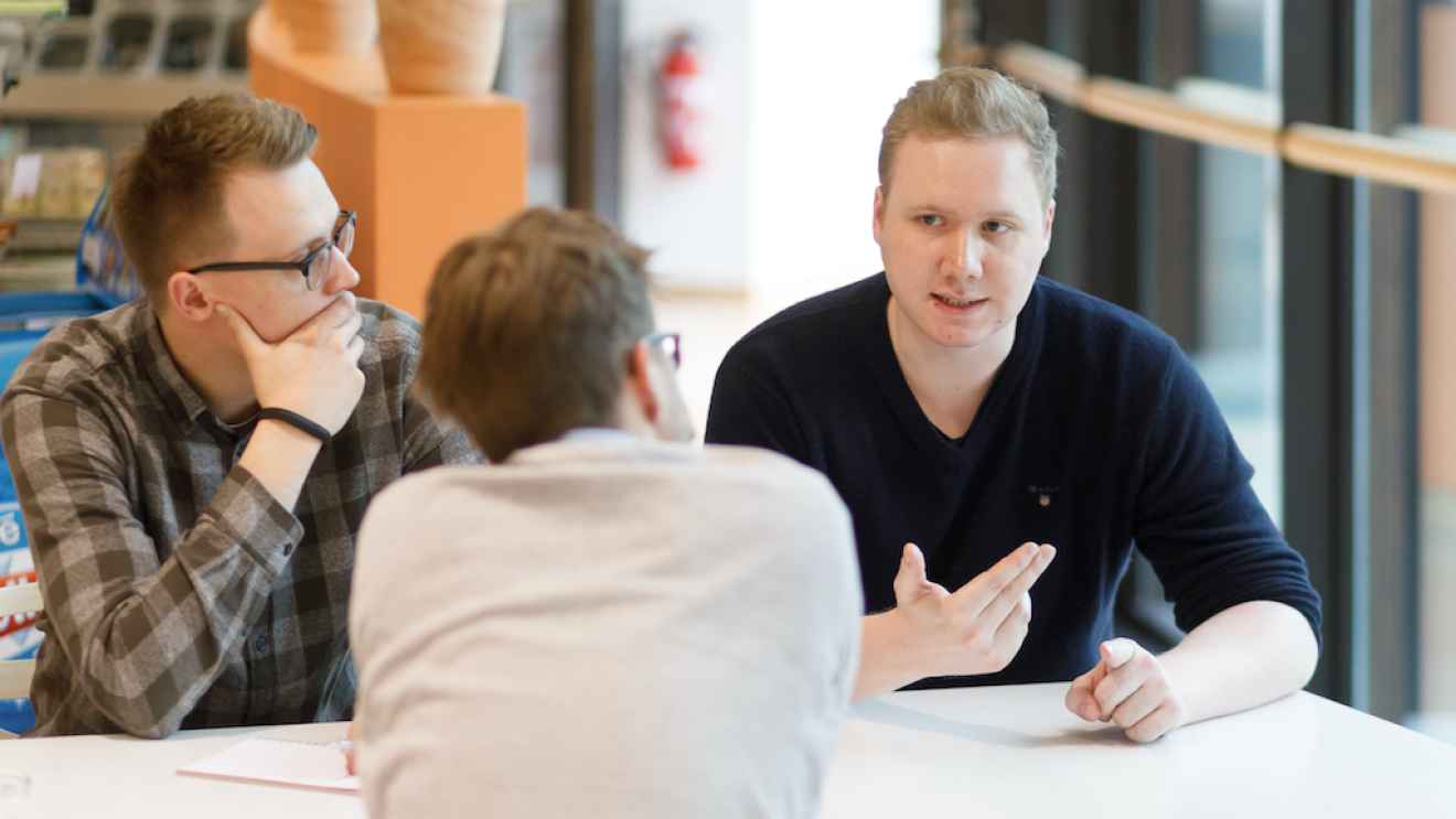 Drei junge Männer sitzen am Tisch und überlegen (zwei sieht man von vorne, einer hat den Rücken zur Kamera gewandt)