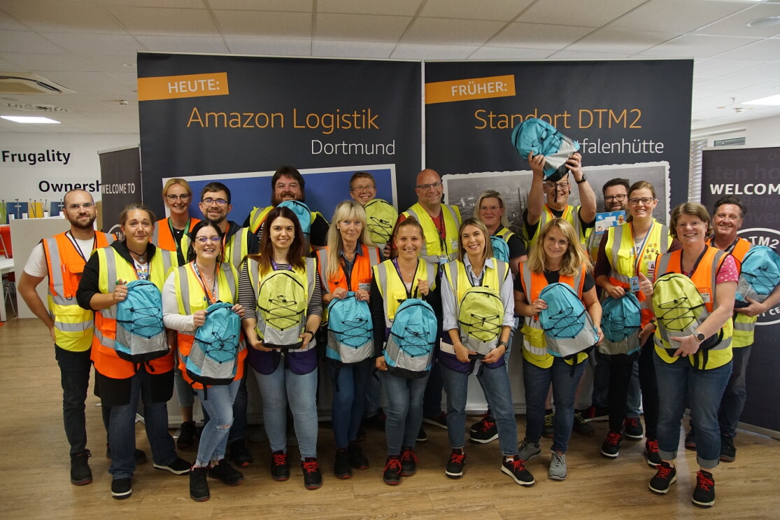 Amazon Mitarbeiterinnen und Mitarbeiter in Sicherheitswesten in Dortmund halten bunte Back to School Rucksäcke in der Hand