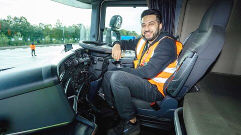 An Amazon delivery driver wears an orange vest and smiles as he sits in the driver seat of a delivery vehicle.