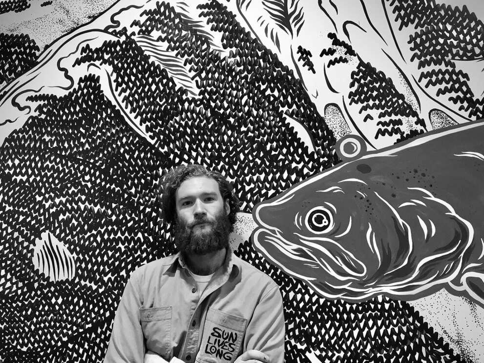 A black and white image of Max Coleman in front of a mural of a large koi fish and rolling hills.