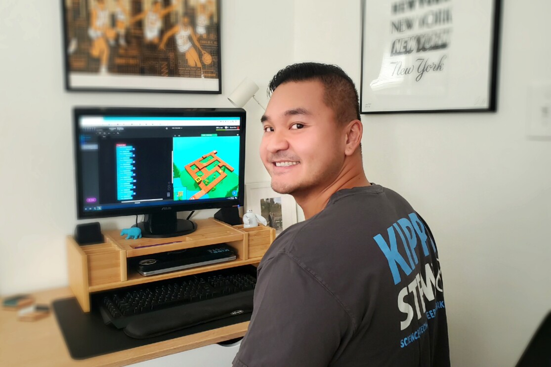 A man sitting at a desk with a computer on it turns around and smiles toward the camera.