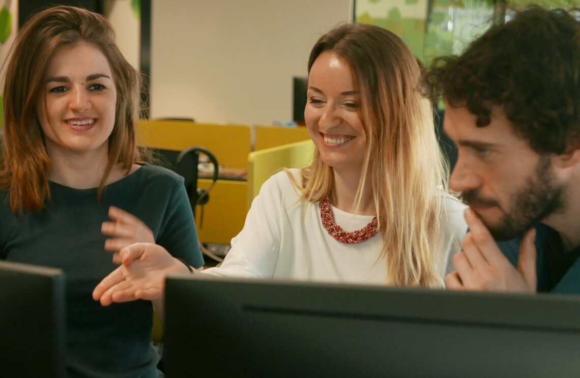 Dos mujeres y un hombre, todos de unos treinta años. Las dos mujeres están sonriendo mirando una pantalla de ordenador y el hombre, con la mano apoyada en su barbilla, observa la pantalla.