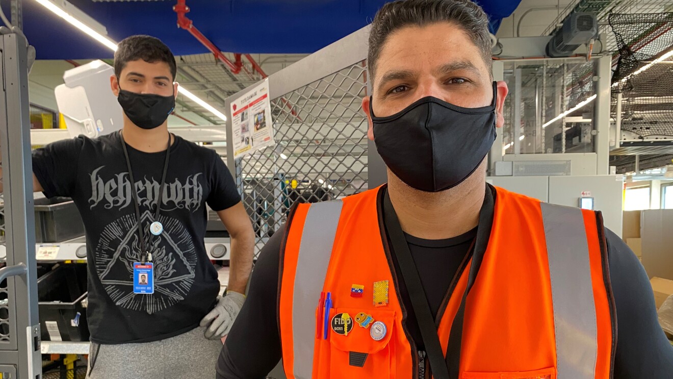 Daniel Fonseca y Diego Fonseca en el centro logístico de Amazon en El Prat de Llobregat (Barcelona). De fondo unas cajas de color negro y el techo con conductos de color azul. En primer plano Daniel con una camiseta térmica estrecha negra de manga larga y un chaleco naranja. Mascarilla de color negro, pelo negro corto y mirando a cámara. En segundo plano está Diego, con un pantalón de deporte una camiseta negra de manga corta. Lleva guantes y la mano izauierda la apoya en la cintura y la derecha en una puerta metálica. Mascarilla negra.