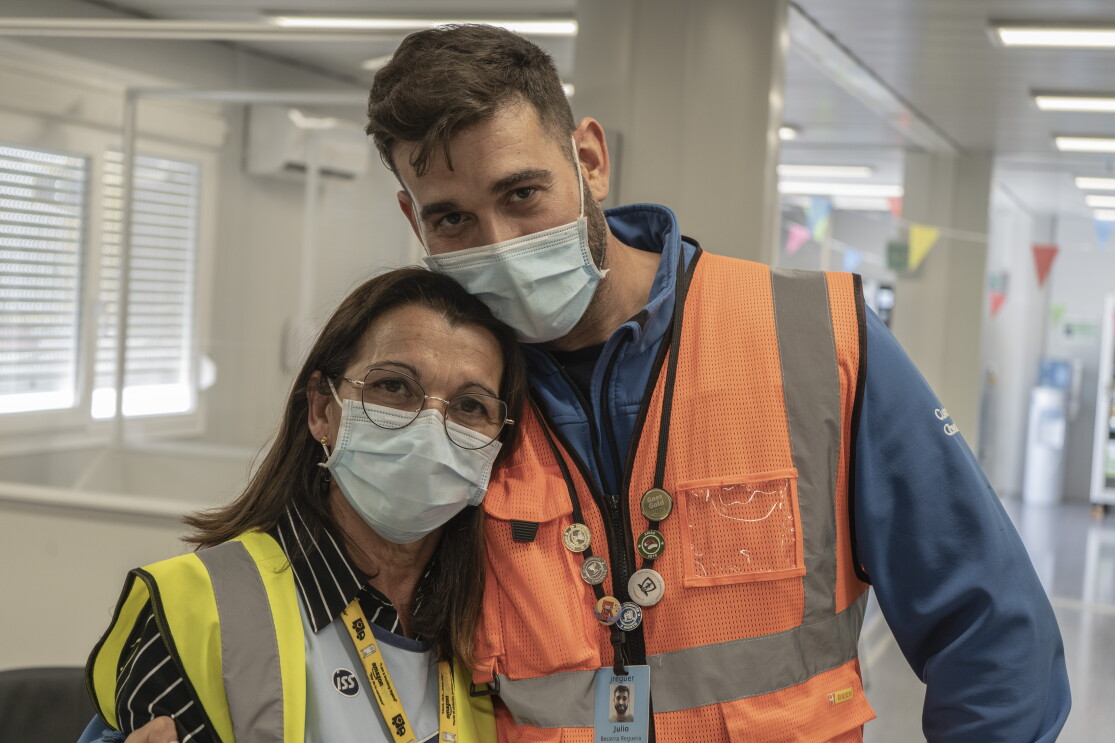María José Requera y Julio Becerra Requera. Situados en las oficinas de la estación logística de Amazon en Málaga. La madre va con un chaleco amarillo, camisa negra con rayas blancas. Lleva gafas, mascarilla quirúrgica y el pelo suelo por el hombro. Su hijo le pasa la cabeza, lleva mascarilla quirúrgica, el pelo corto, un forro polar azul y un chaleco naranja. 