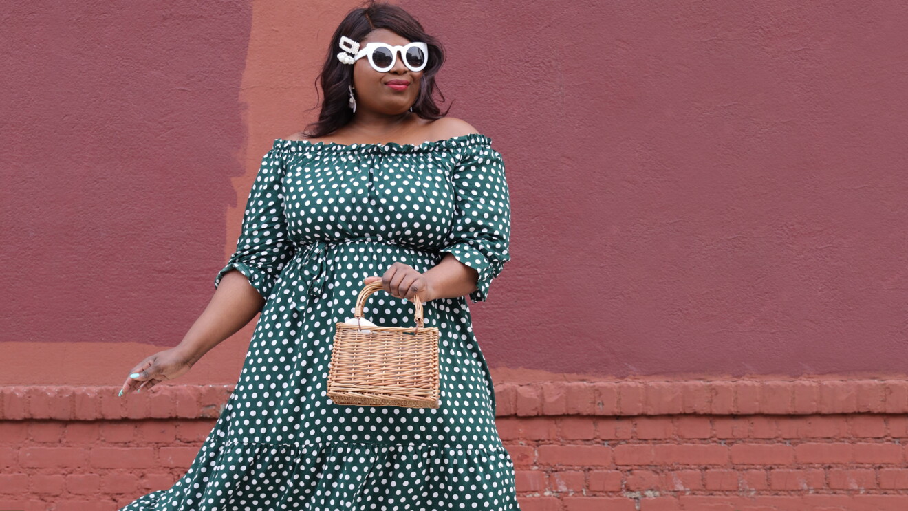 A stylish woman in front of a brick and stucco wall wears an off-the-shoulder dress with white polka dots, white retro sunglasses, and carries a small wicker purse.