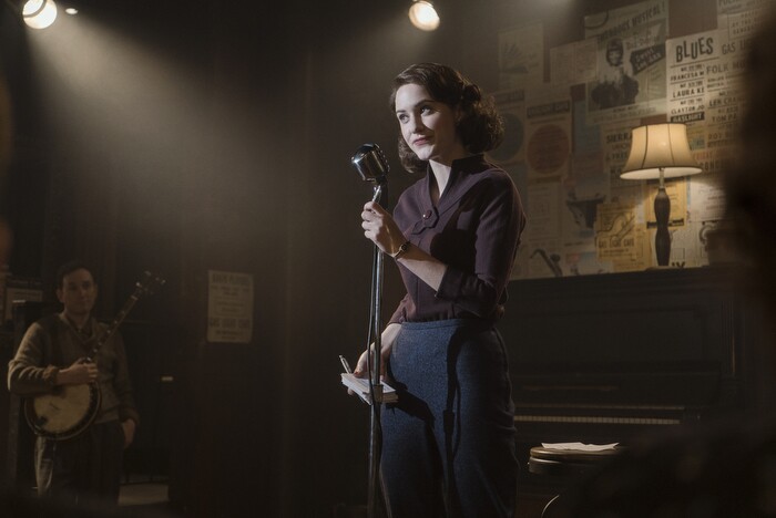 An image of Mrs. Maisel standing on a stage with a notepad and pen in her hand and a microphone in front of her. She is wearing a dark purple top and dark blue pants reflecting the "downtown" style.