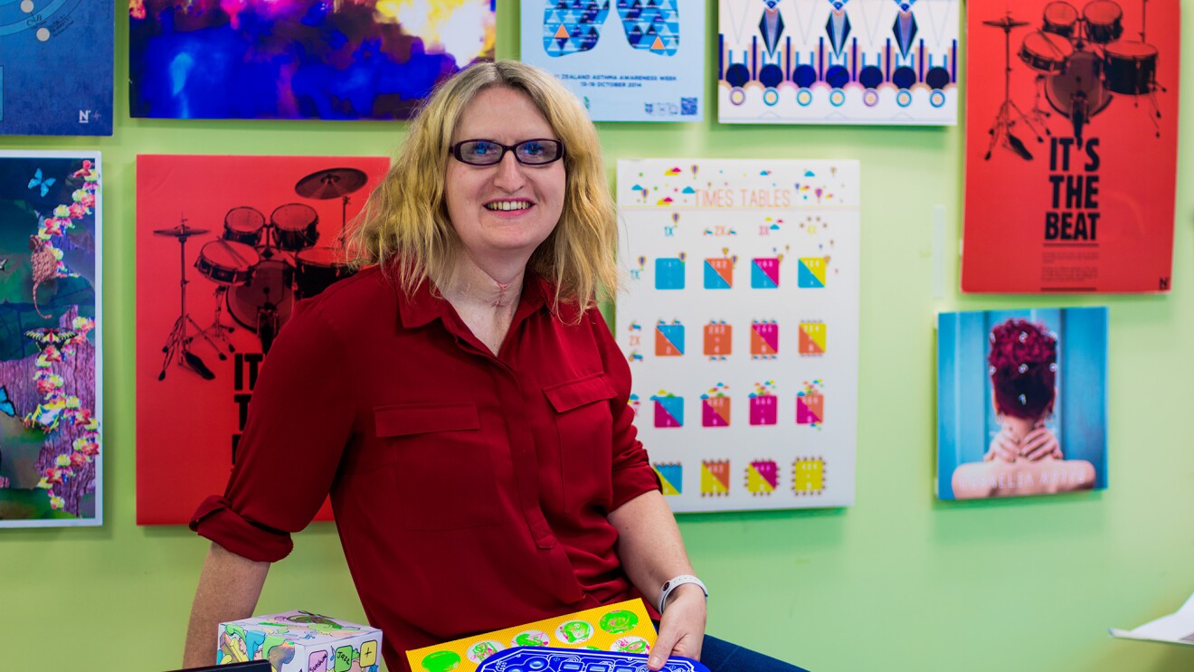 A blonde woman wearing glasses, jeans, and red button up shirt is seated on a tabletop in a room painted green, with myriad artwork hung on the wall.