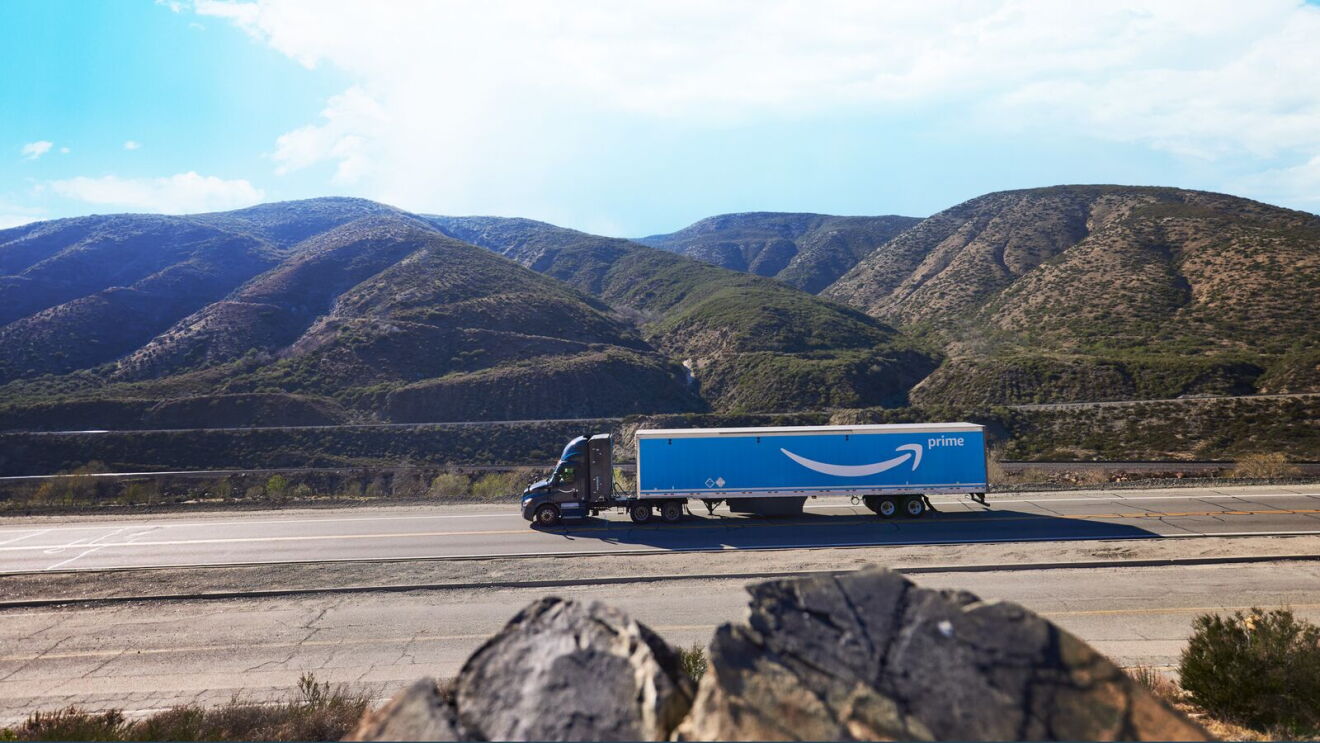 An Amazon delivery truck on the road with mountains in the background.