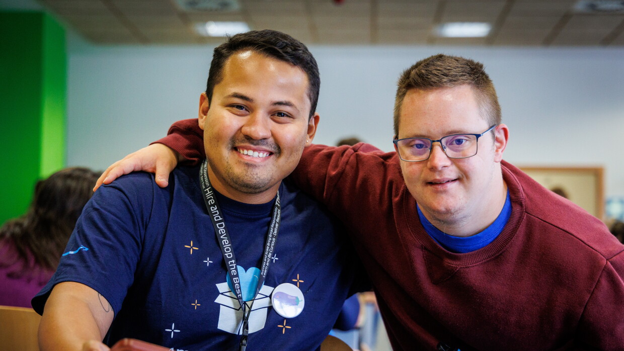 En las instalaciones de Down Madrid en Madrid aparecen 2 personas en primer plano. Un trabajador de Amazon y un participante de la fundación . El trabajador de Amazon van con la misma camiseta azul  y el badge colgado y el de Down Madrid con deportivas con un jersey de color morado. La camiseta del trabajador de Amazon lleva un dibujo que es una caja de Amazon de la que sale un corazón y debajo las palabras Amazon Volunteer. 