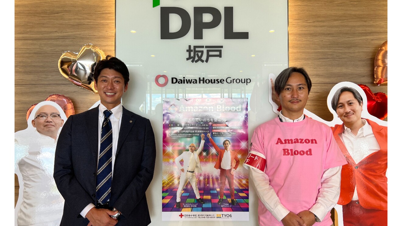 物流拠点のロゴとAmazon Bloodのポスターの前で笑顔の村上さんと松下さんの写真