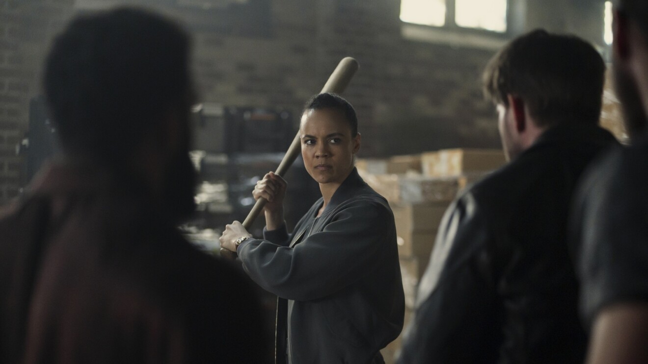 Still from 'Neagley' of woman in dark jacket holding baseball bat in defensive stance while confronting two people in warehouse