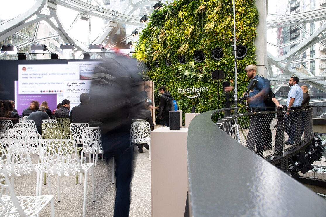 A man walking into the September 2018 Devices announcement event. To the left are chairs in front of a large screen. To the right others stand against a railing. Behind the scene is a living wall in The Spheres.