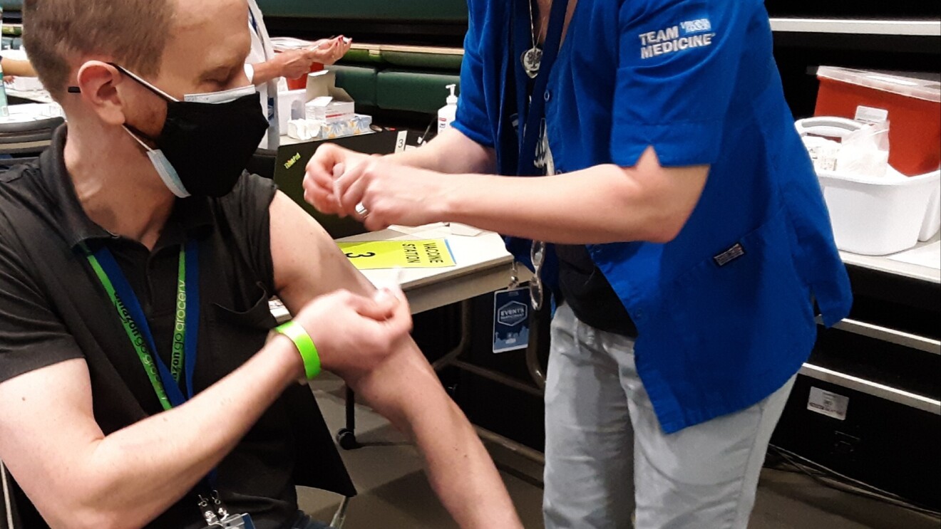 A man rolls up his sleeve while a medical professional administers the COVID-19 vaccine in his arm. Bot individuals are wearing masks.