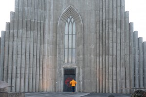 An image of a person wearing a yellow coat in front of two doors on a tall grey wall. 