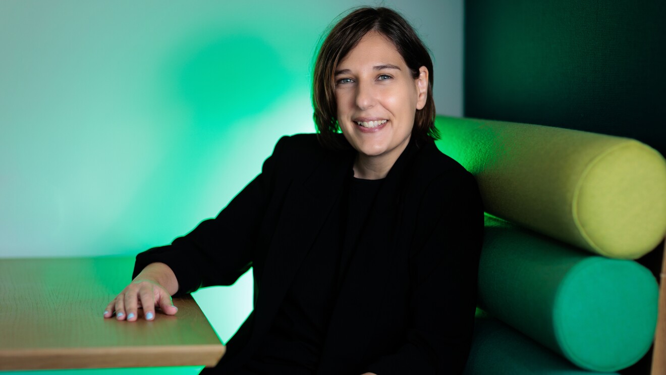 María Alonso vestida con un traje pantalón de color negro, con un jersey negro también debajo de la americana. Con el pelo corto y los ojos azules. Las manos con las uñas azules. Con la mano derecha apoyada en la mesa y la otra en la pierna. Sentada en un sofá con tonos verdes y luces verdes de fondo.
