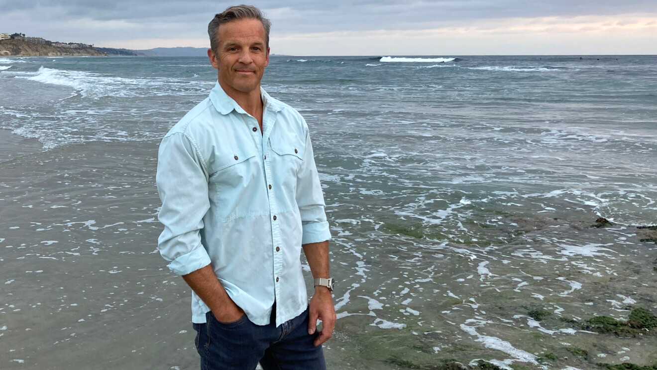 Author Dan Sheehan stands on an ocean beach wearing jeans and a button-down shirt.