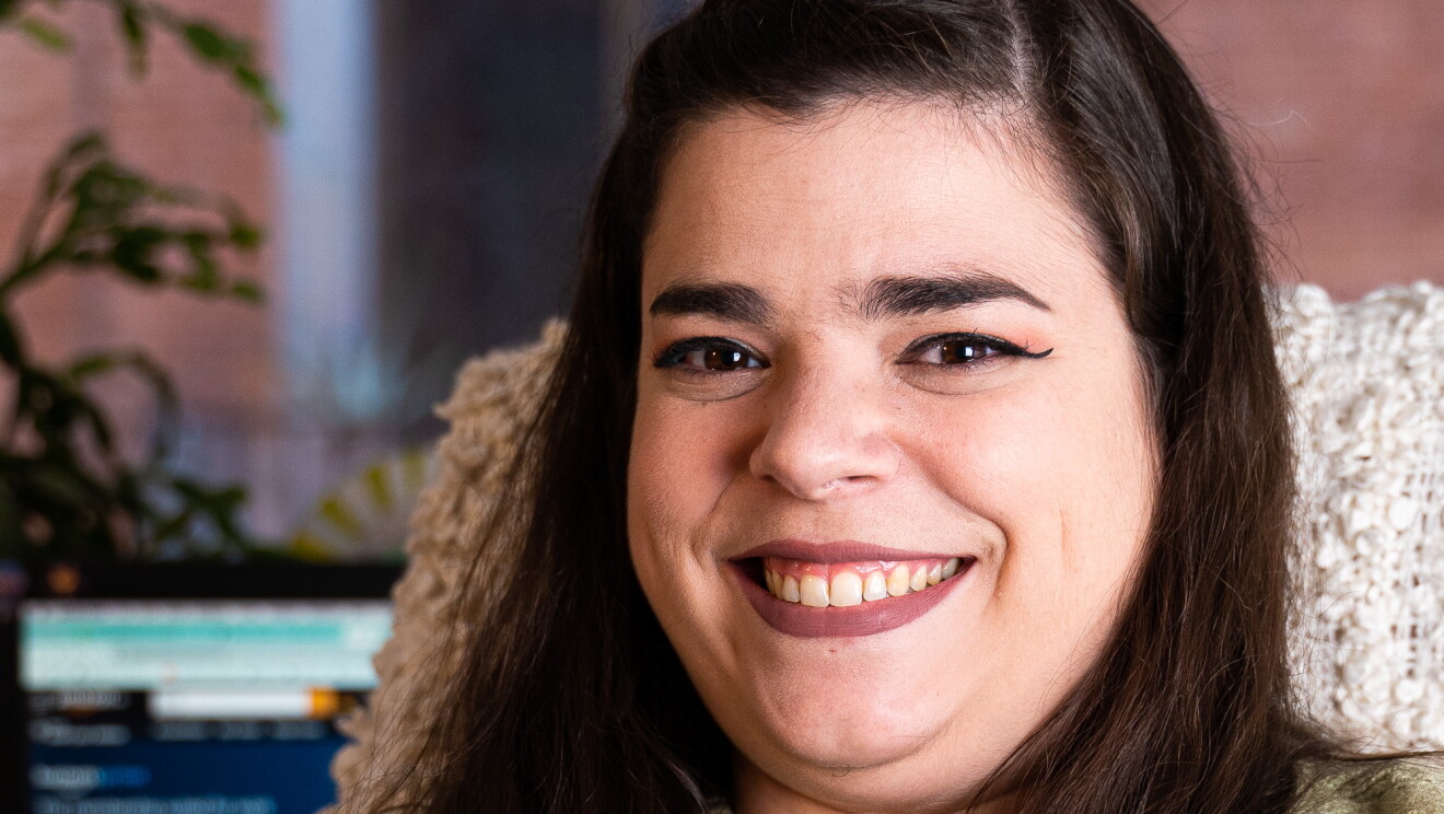 Violeta González. Mirando a cámara sonriente con el pelo oscuro hasta el pecho. Cejas pronunciadas y labios maquillados. Jersey de punto verde claro. De fondo una silla con una funda de punto y una mesa con libros.  