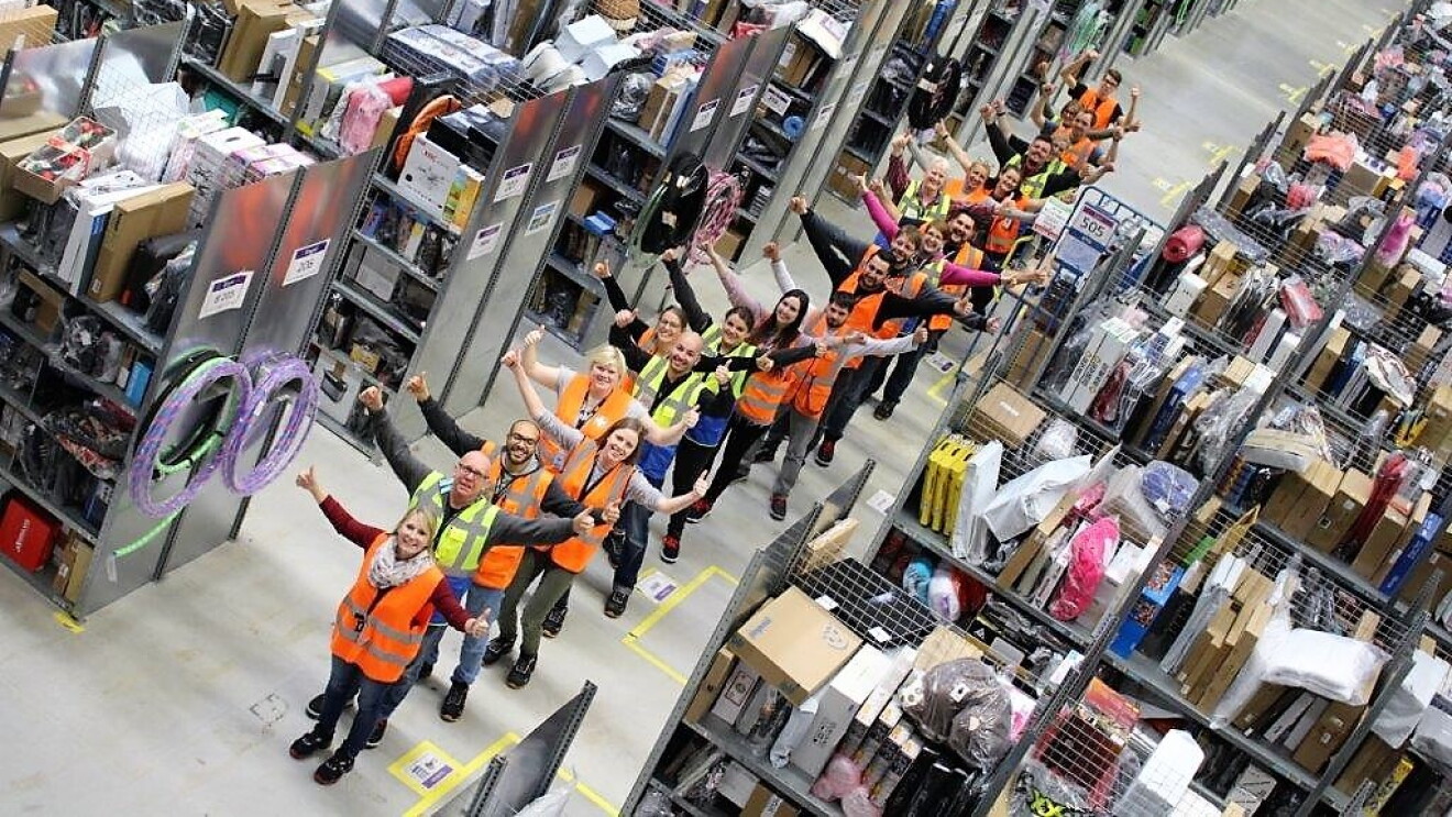Eine Reihe von Logistikmitarbeitern stehen nacheinander im einem Regalgang, die Daumen nach oben