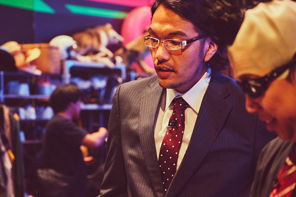 さまざまな小道具が積まれた棚を背景に立つ2人の男性のクローズアップ。左側の男性は眼鏡に縦じまのスーツでビジネスマン風、赤いネクタイ。右側の男性はモヒカンのかつらとサングラスを着用。