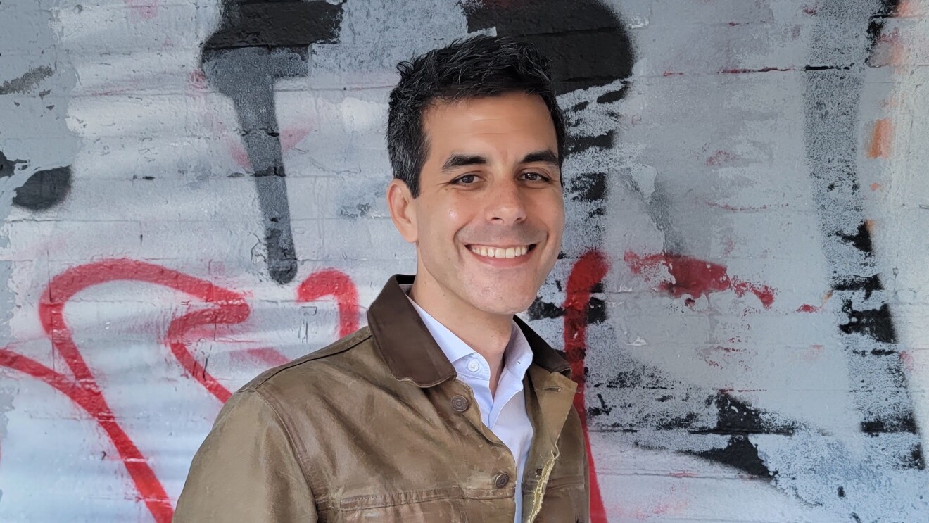 Adam Siegel smiles wearing a tan jacket and stands in front of a graffiti wall.