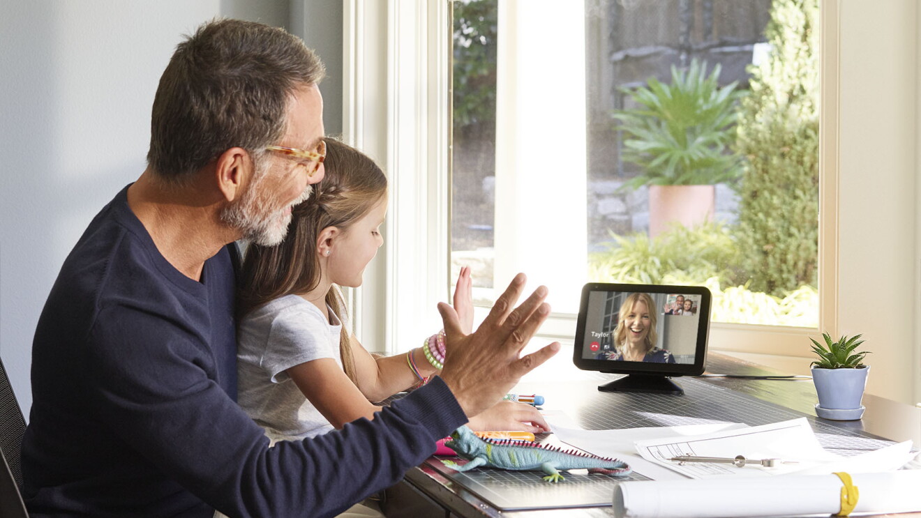 A little girl sits on a man's lap at a desk. They are both waving and looking at an Echo Show device, on which a woman is smiling and engaging with them. 
